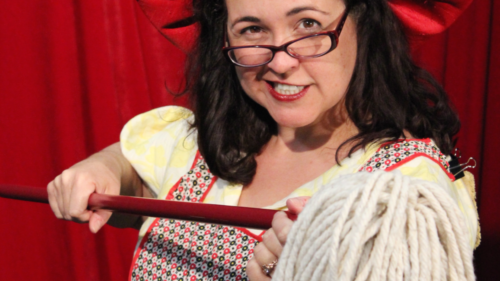 A white woman with brown hair and glasses points a mop at the camera.