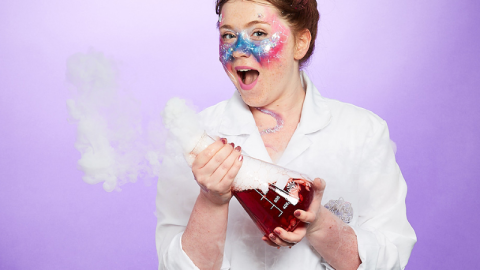 A white woman in a lab coat, with glitter on her face and in her hair, holds a bubbling scientific flask with an excited smile.