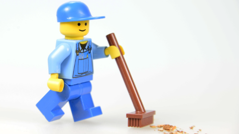 A Lego minifigure dressed in blue work clothes and a hat pushes a plastic broom.