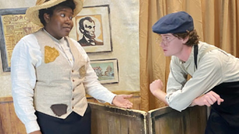 A Black woman and a white man, both in pre-Civil War era clothing, stand talking in front of a minimalist theatrical set.