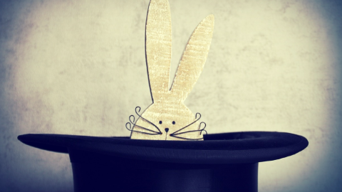 A white wooden rabbit with thin metal whiskers peeks out of a black magician's hat.