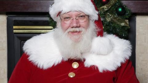 A smiling man with a white full beard, wearing wire-rimmed glasses and a traditional red-and-white Santa suit.