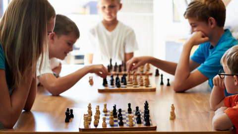 A diverse group of children play chess at a wooden table.