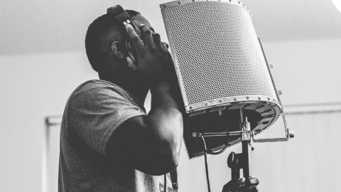 Black-and-white image of a Black man in a short-sleeved shirt singing into a professional microphone, wearing headphones.