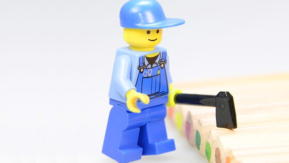 A Lego minifigure in blue coveralls chops at a row of colored pencils with a tiny axe.