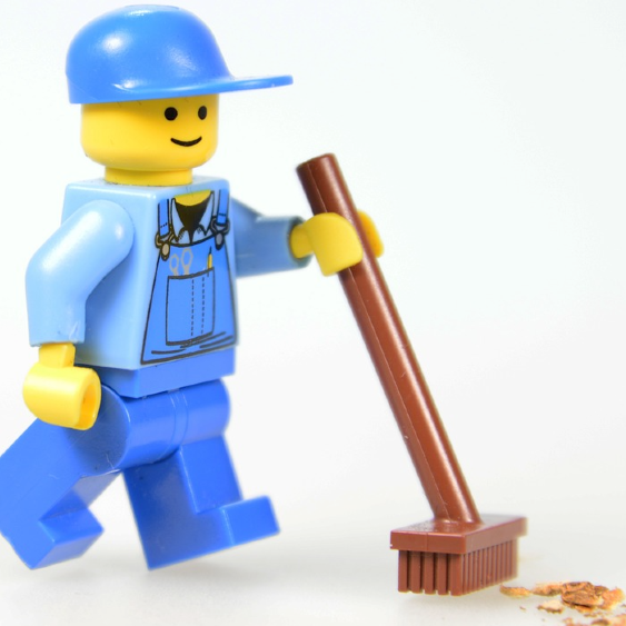 A Lego minifigure dressed in blue work clothes and a hat pushes a plastic broom.